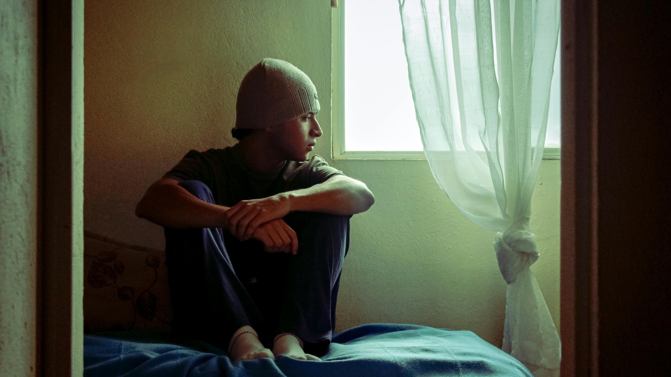 Adolescente en actitud melancólica, mirando por la ventana de una habitación con iluminación natural tenue.