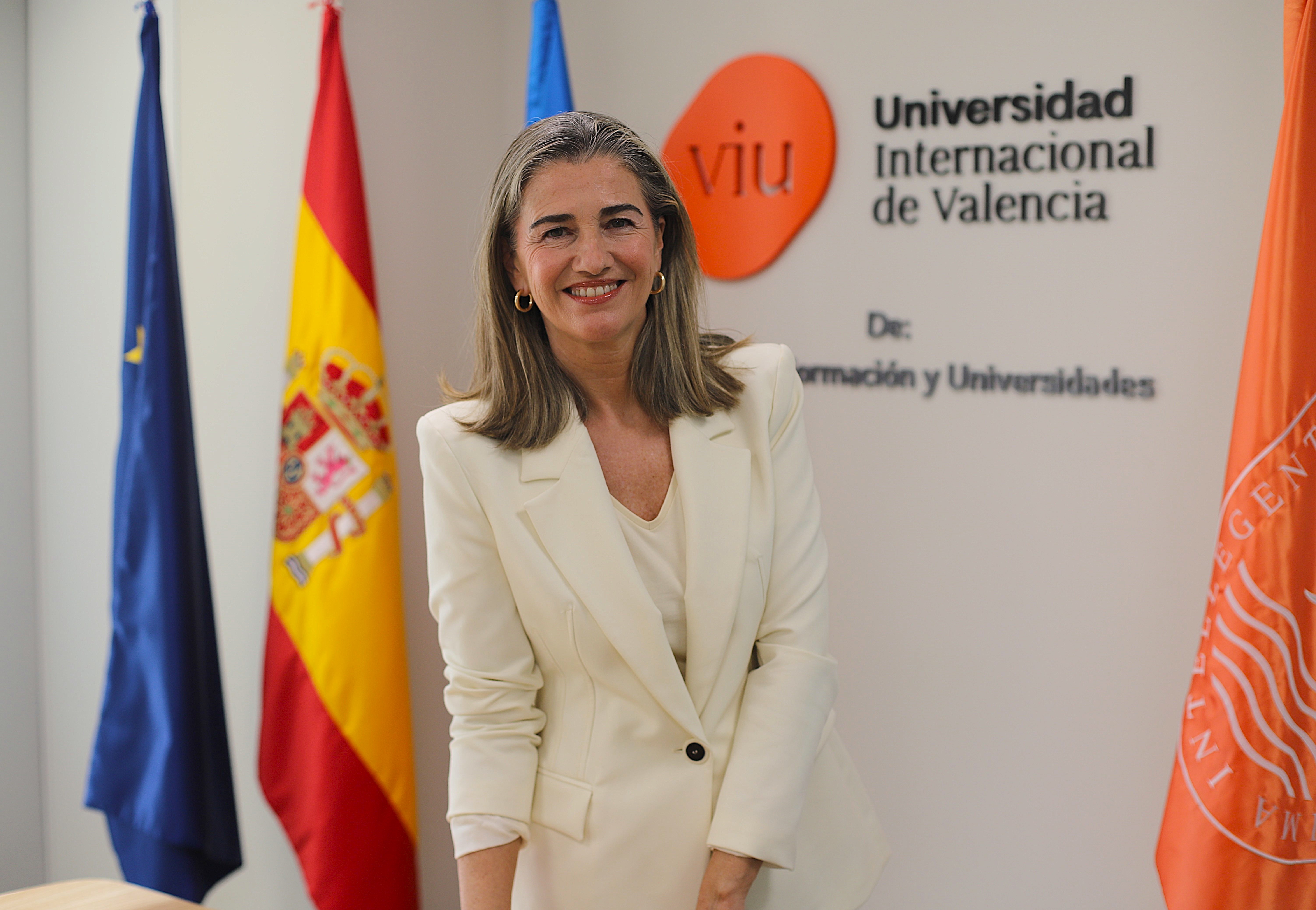 Retrato de la Dra. Julia Martínez, decana de la Facultad de Ciencias Sociales y Jurídicas de VIU. Viste de blanco, mira sonriente a cámara y de fondo tiene el logo de VIU y banderas institucionales