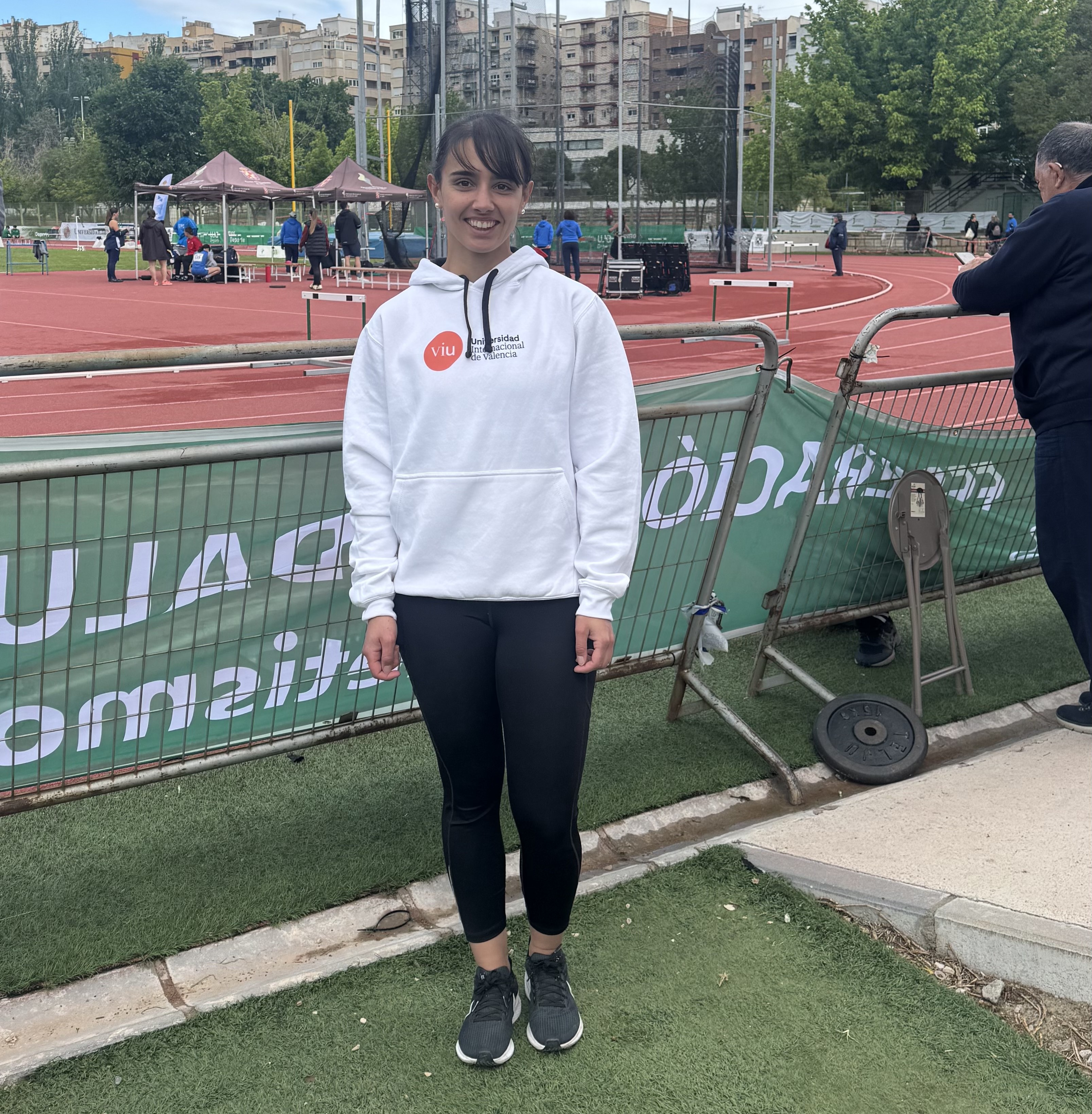 Lydia Cuesta con sudadera blanca con el logo de VIU en naranja, posando de pie con la pista de atletismo de fondo