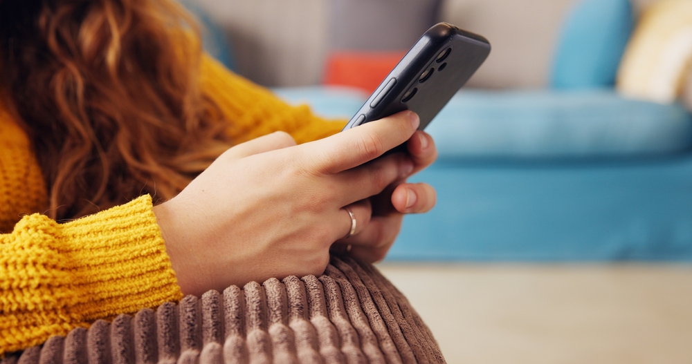 Persona interactuando con un dispositivo móvil y la inteligencia artificial Primer plano de las manos de una mujer usando un teléfono móvil, apoyada en un cojín de color marrón, relacionando la tecnología digital con la empatía y la inteligencia artificial.
