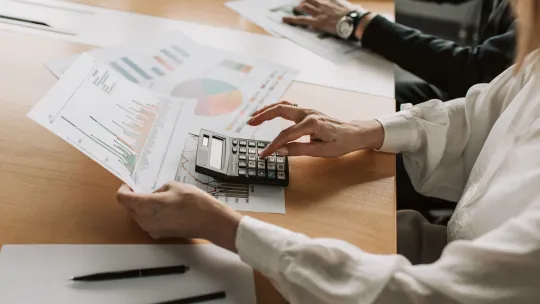 Análisis de documentos y métricas financieras en las Big Four Persona en una mesa de oficina analizando informes financieros impresos con gráficos y utilizando una calculadora.