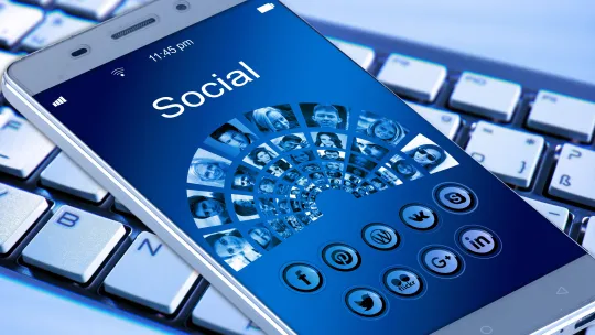 Dispositivo móvil con interfaz de comunicación social sobre teclado eléfono móvil sobre un teclado de ordenador mostrando en pantalla una interfaz azul con la palabra Social, rodeada de iconos de redes sociales y retratos de personas conectados en red.