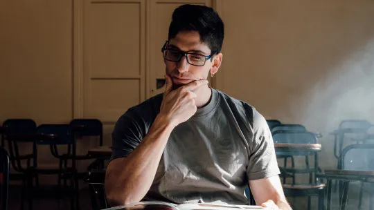 Estudiante en aula analizando las diferencias entre un MBA y un máster Hombre joven con gafas y camiseta gris reflexivo, sosteniendo su mentón mientras revisa un libro abierto en un aula universitaria iluminada por luz natural, evaluando la diferencia entre un MBA y un máster.