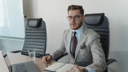Director comercial en reunión estratégica de trabajo Hombre profesional con traje claro, gafas y corbata, sentado en una sala de reuniones frente a un ordenador portátil con un cuaderno abierto sobre la mesa.