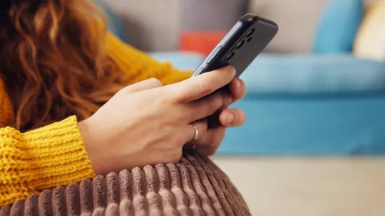 Persona interactuando con un dispositivo móvil y la inteligencia artificial Primer plano de las manos de una mujer usando un teléfono móvil, apoyada en un cojín de color marrón, relacionando la tecnología digital con la empatía y la inteligencia artificial.