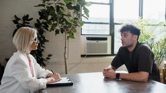 Una psicóloga clínica profesional con gafas y chaqueta blanca toma notas mientras mantiene una conversación terapéutica con un paciente joven en una oficina iluminada.