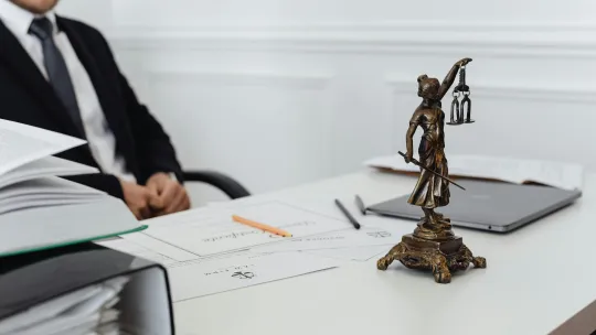 Escena de despacho jurídico con la figura de Themis y documentación legal preparada para el examen de acceso a la abogacía Estatua dorada de la Justicia sobre una mesa de despacho junto a documentos legales, un portátil y útiles de escritura, con un abogado de traje oscuro al fondo