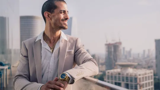 Hombre joven con traje claro y reloj de pulsera, apoyado en una barandilla de cristal con un paisaje urbano de edificios modernos al fondo, proyectando una actitud de liderazgo y visión.