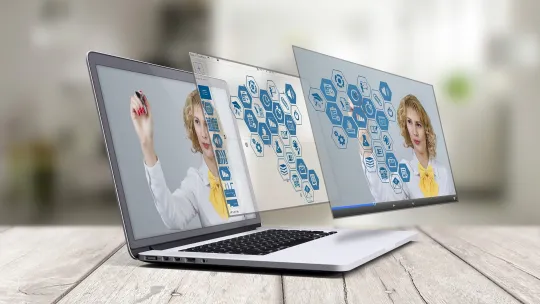 Ordenador portátil sobre una mesa de madera mostrando una interfaz virtual con iconos educativos mientras una profesora interactúa con el contenido en pantalla.