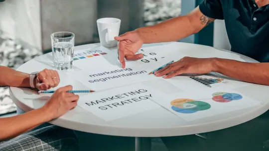 Profesionales revisando una estrategia con herramientas de marketing automation Dos profesionales reunidos alrededor de una mesa blanca analizando documentos sobre estrategias y segmentación, representando el análisis para elegir herramientas de marketing automation.