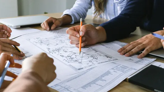 Equipo de trabajo analizando planos de ingeniería en organización industrial Un grupo de personas trabajando alrededor de una mesa de madera, revisando planos técnicos y esquemas de ingeniería en organización industrial con un lápiz.