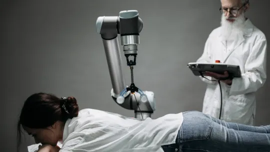 Asistencia robótica e inteligencia artificial en el ámbito sanitario Paciente tumbada en una camilla recibiendo terapia asistida por un brazo robótico médico, mientras un profesional sanitario revisa los datos en una tablet.