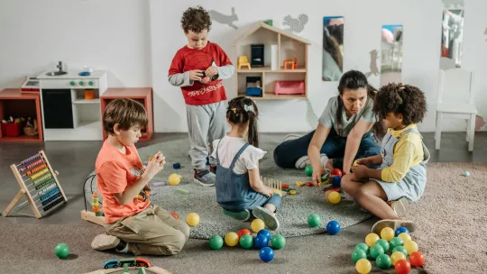 Niños jugando con pelotas y juguetes de madera junto a su profesora en un entorno de juego inclusivo para niños.