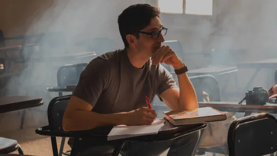 Estudiante en un aula analizando las menciones en Educación Primaria Hombre joven con gafas y camiseta gris sentado en un pupitre de aula, sosteniendo un bolígrafo sobre un cuaderno con actitud reflexiva.