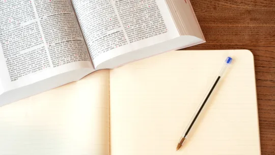 Material de estudio para oposiciones de derecho Libro de texto jurídico abierto sobre una mesa de madera junto a un cuaderno en blanco y un bolígrafo azul, simbolizando el estudio para las oposiciones de derecho.