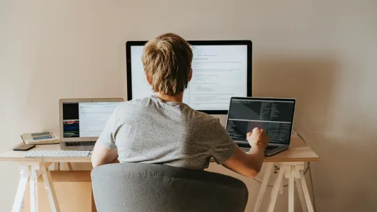 Estudiante de Máster en Inteligencia Artificial Online en su entorno de trabajo Un joven estudiante trabaja en un escritorio con un monitor grande y dos portátiles abiertos. En las pantallas se ven líneas de código y material de estudio online de Inteligencia Artificial.