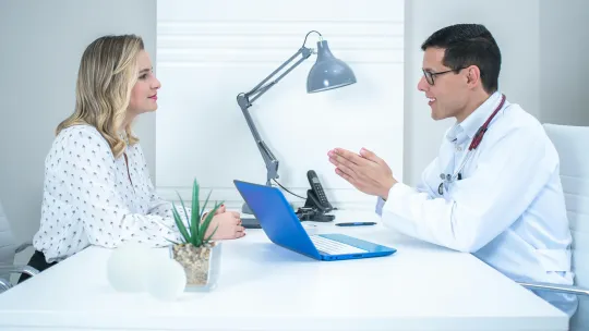 Un neuropsicólogo clínico con bata blanca explica un diagnóstico a una paciente frente a un ordenador portátil azul en una consulta