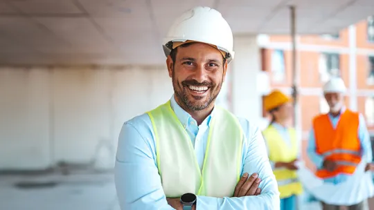 Responsable de seguridad y salud supervisando una obra de construcción Hombre sonriente en primer plano con casco blanco de protección, camisa clara y chaleco reflectante amarillo con los brazos cruzados, mientras al fondo en el espacio de trabajo de la obra interactúan otros profesionales con cascos y chalecos.