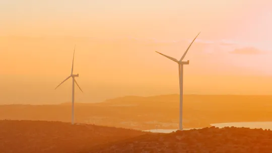 Parque eólico en el sector energético al atardecer Dos aerogeneradores eólicos situados en una colina frente al mar durante un atardecer en tonos cálidos anaranjados y dorados.
