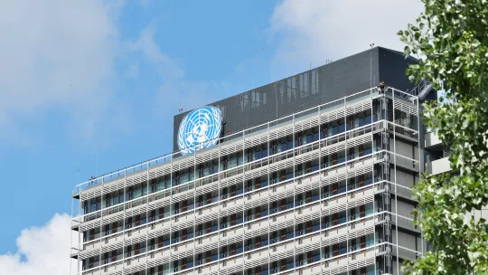Edificio institucional de Naciones Unidas Vista exterior de la fachada de un edificio institucional de la ONU con el emblema de Naciones Unidas visible en la parte superior, bajo un cielo parcialmente nublado.