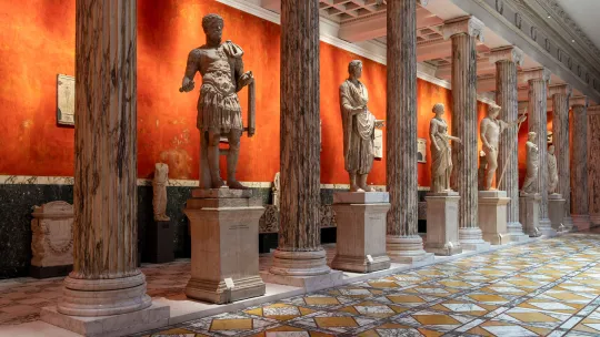 Galería de arte clásico para trabajar en museos Galería de museo de arte clásico con estatuas de mármol expuestas sobre pedestales entre columnas de mármol veteado, ante una pared naranja y un suelo geométrico.
