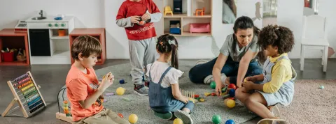 Niños jugando con pelotas y juguetes de madera junto a su profesora en un entorno de juego inclusivo para niños.