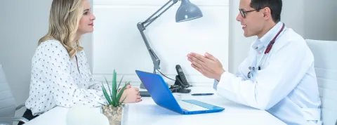 Un neuropsicólogo clínico con bata blanca explica un diagnóstico a una paciente frente a un ordenador portátil azul en una consulta