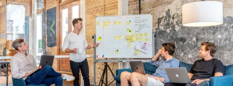 cuatro-hombres-trabajando-en-una-sala-tres-sentados-con-portatiles-y-uno-de-pie-los-cuatro-mirando-una-pizarra-donde-estan-tratando-el-tema-de-las-sociedades-de-capital