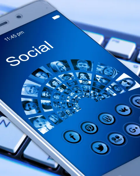 eléfono móvil sobre un teclado de ordenador mostrando en pantalla una interfaz azul con la palabra Social, rodeada de iconos de redes sociales y retratos de personas conectados en red.