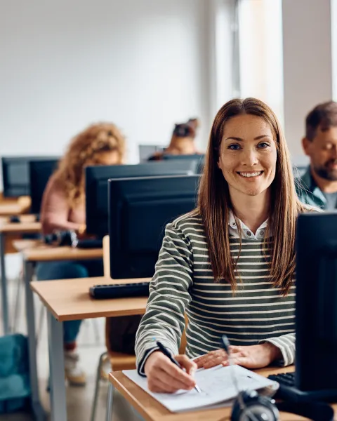 Estudiante sonriente sentada ante un ordenador en un aula informática, tomando notas durante una sesión donde se dan clases de FP.