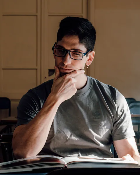 Hombre joven con gafas y camiseta gris reflexivo, sosteniendo su mentón mientras revisa un libro abierto en un aula universitaria iluminada por luz natural, evaluando la diferencia entre un MBA y un máster.