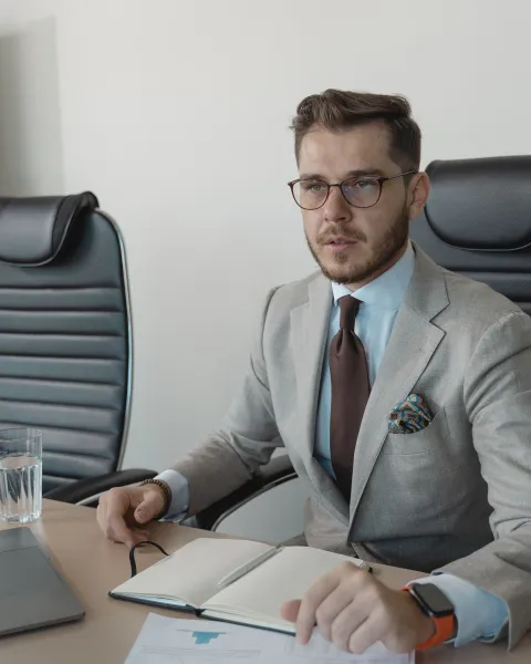 Hombre profesional con traje claro, gafas y corbata, sentado en una sala de reuniones frente a un ordenador portátil con un cuaderno abierto sobre la mesa.