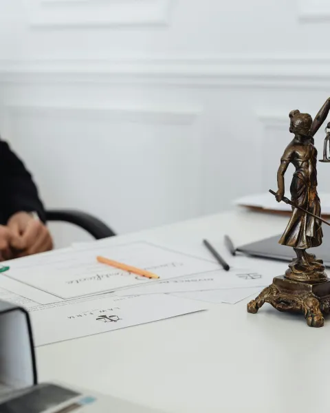 Estatua dorada de la Justicia sobre una mesa de despacho junto a documentos legales, un portátil y útiles de escritura, con un abogado de traje oscuro al fondo