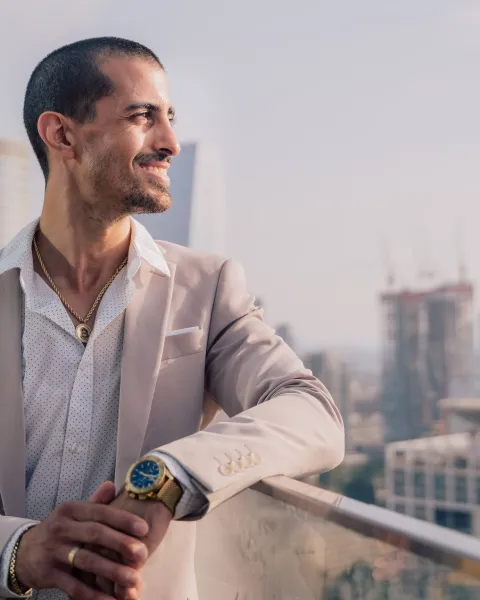 Hombre joven con traje claro y reloj de pulsera, apoyado en una barandilla de cristal con un paisaje urbano de edificios modernos al fondo, proyectando una actitud de liderazgo y visión.