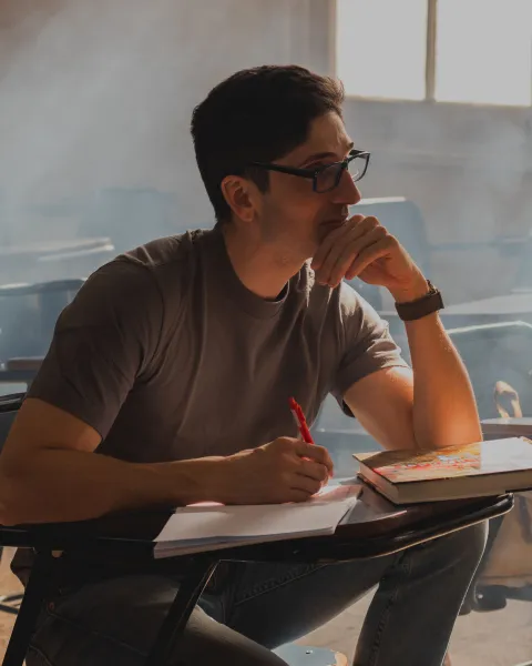 Hombre joven con gafas y camiseta gris sentado en un pupitre de aula, sosteniendo un bolígrafo sobre un cuaderno con actitud reflexiva.