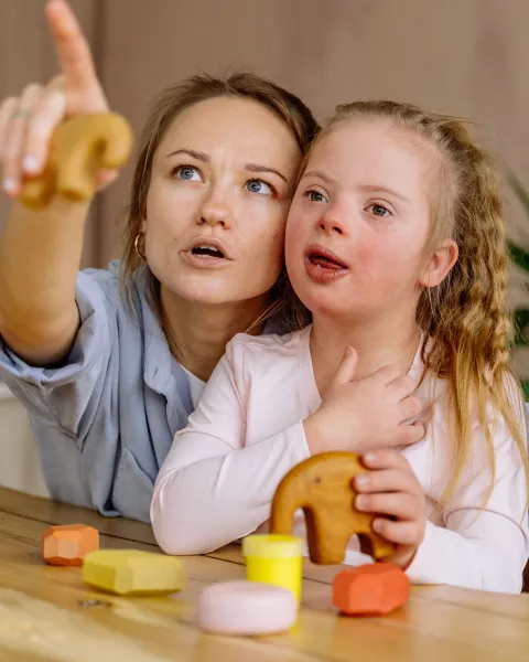 Profesora o educadora señalando hacia arriba mientras acompaña a una niña con síndrome de Down en una mesa con juguetes de madera coloridos, en un entorno de aprendizaje enfocado en necesidades educativas especiales.