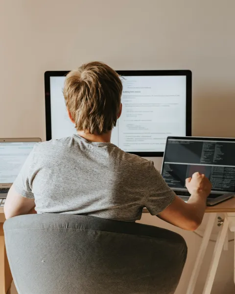 Un joven estudiante trabaja en un escritorio con un monitor grande y dos portátiles abiertos. En las pantallas se ven líneas de código y material de estudio online de Inteligencia Artificial.
