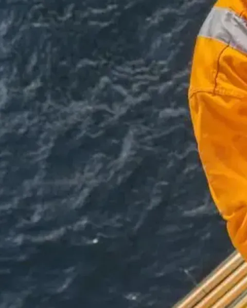 Trabajador industrial con mono naranja, arnés de seguridad y casco amarillo realizando labores en altura sobre el mar.