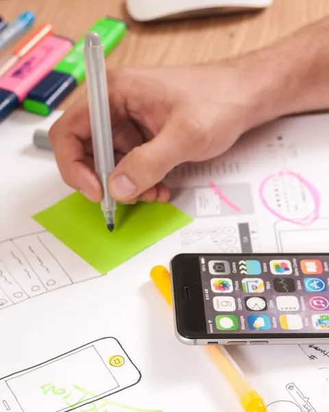 mano-de-una-persona-que-esta-trabajando-en-un-proceso-tecnologico-y-va-a-apuntar-en-un-posit-que-esta-en-una-mesa-con-papeles-un-movil-y-subrayadores