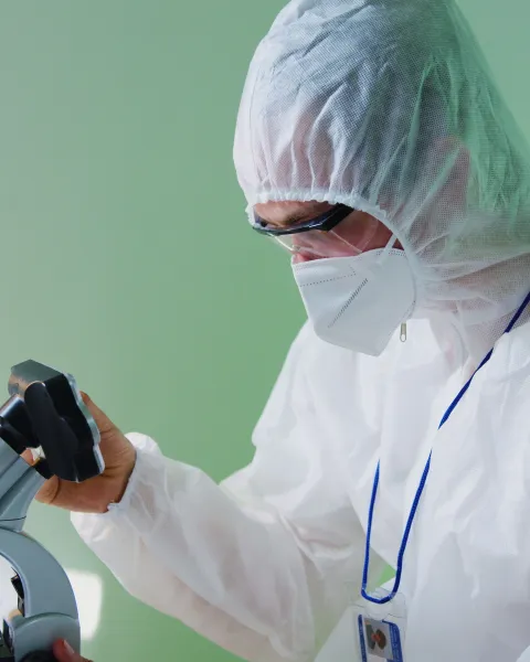 Científico con traje de protección biológica blanco, mascarilla y gafas de seguridad observando una muestra a través de un microscopio en un laboratorio.