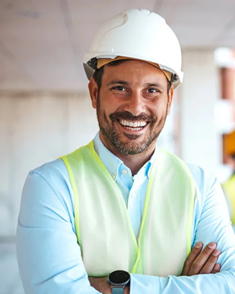 Hombre sonriente en primer plano con casco blanco de protección, camisa clara y chaleco reflectante amarillo con los brazos cruzados, mientras al fondo en el espacio de trabajo de la obra interactúan otros profesionales con cascos y chalecos.