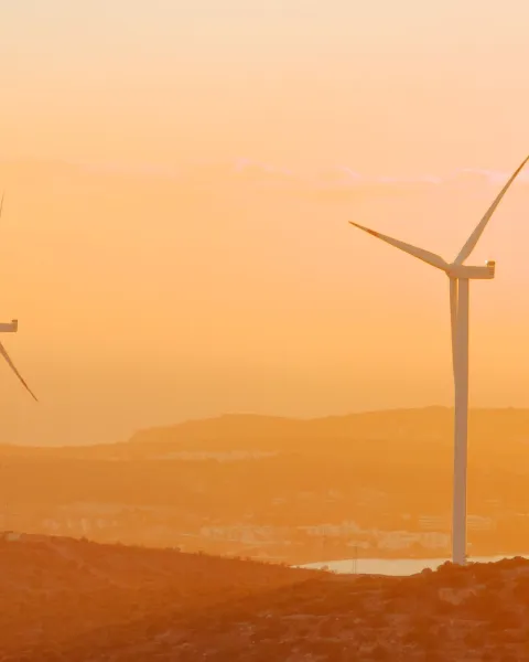 Dos aerogeneradores eólicos situados en una colina frente al mar durante un atardecer en tonos cálidos anaranjados y dorados.