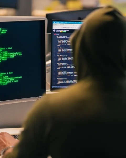 Persona de espaldas con capucha oscura frente a múltiples monitores que muestran líneas de código informático en verde sobre fondo negro, ilustrando los tipos de ciberataques y la ciberseguridad.