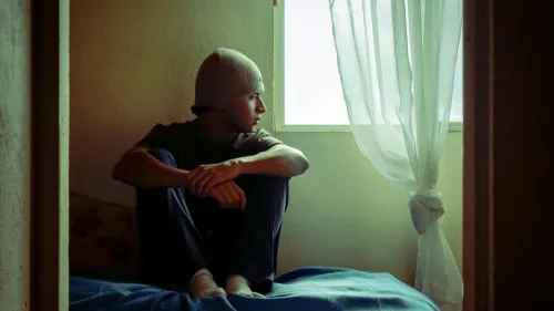 Adolescente en actitud melancólica, mirando por la ventana de una habitación con iluminación natural tenue.