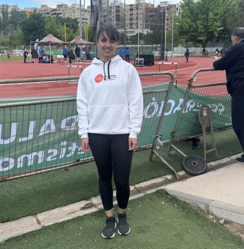 Lydia Cuesta con sudadera blanca con el logo de VIU en naranja, posando de pie con la pista de atletismo de fondo