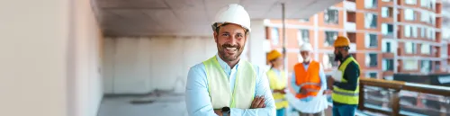 Hombre sonriente en primer plano con casco blanco de protección, camisa clara y chaleco reflectante amarillo con los brazos cruzados, mientras al fondo en el espacio de trabajo de la obra interactúan otros profesionales con cascos y chalecos.